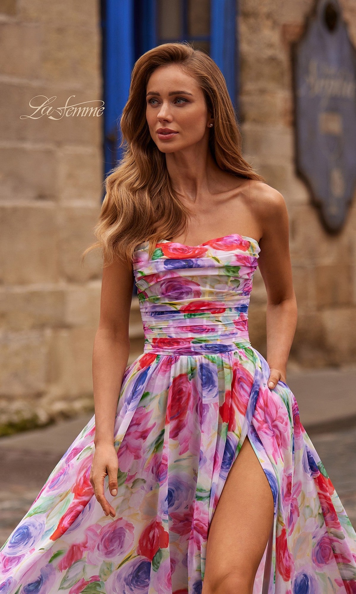 A woman stands outdoors by a stone wall and blue door, wearing the La Femme 33487 long prom dress featuring a strapless multicolor floral print, ruched fitted bodice, and high slit.